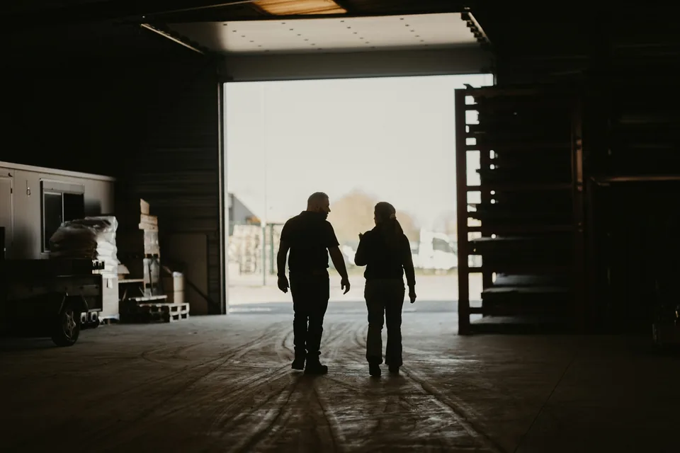 Two people in silhouette walk side by side across a dark warehouse toward its open bay door.