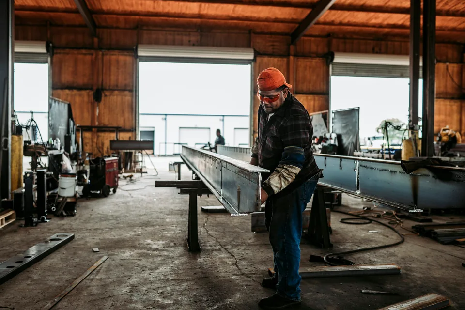 A welder in a hard hat and safety glasses checks the end of a long steel beam in a fabrication shop, its bay doors open behind him.