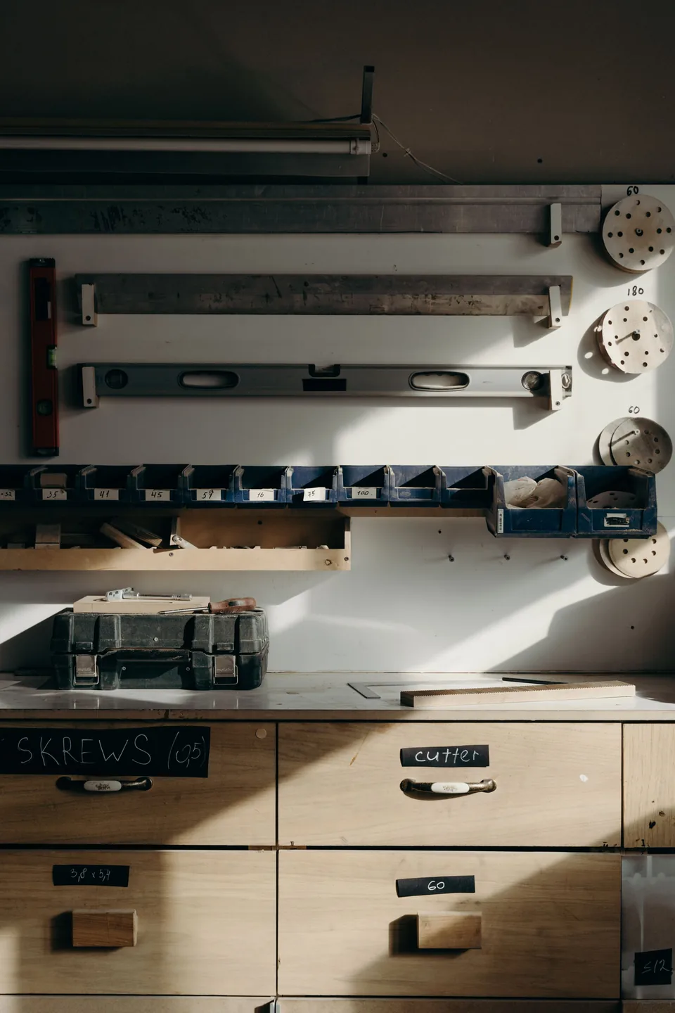 A tidy workshop wall: levels and rules hung on a white board, a row of numbered bins, a toolbox on the bench and labelled drawers below.