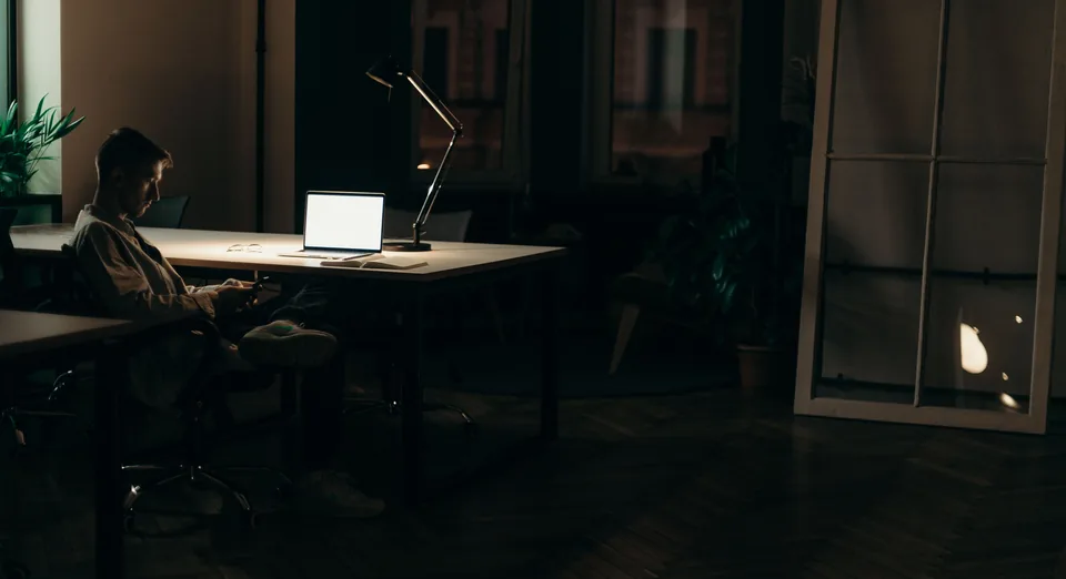 A man working late at a laptop under a desk lamp, alone in a dark loft office.