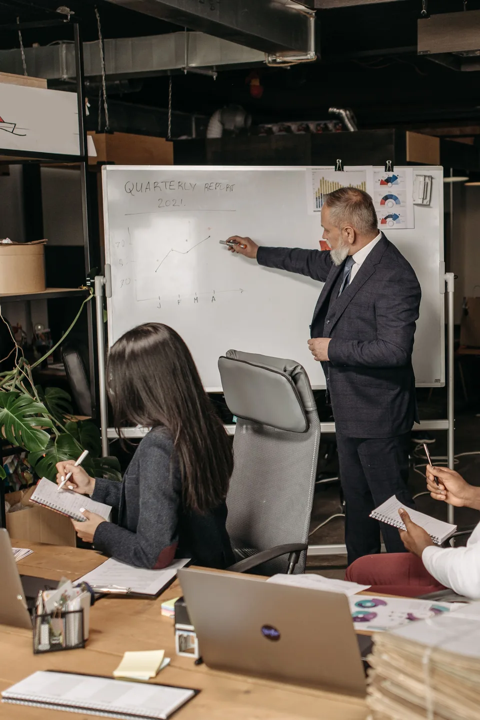 A consultant in a suit charts a quarterly report on a whiteboard while the people at the table take notes.