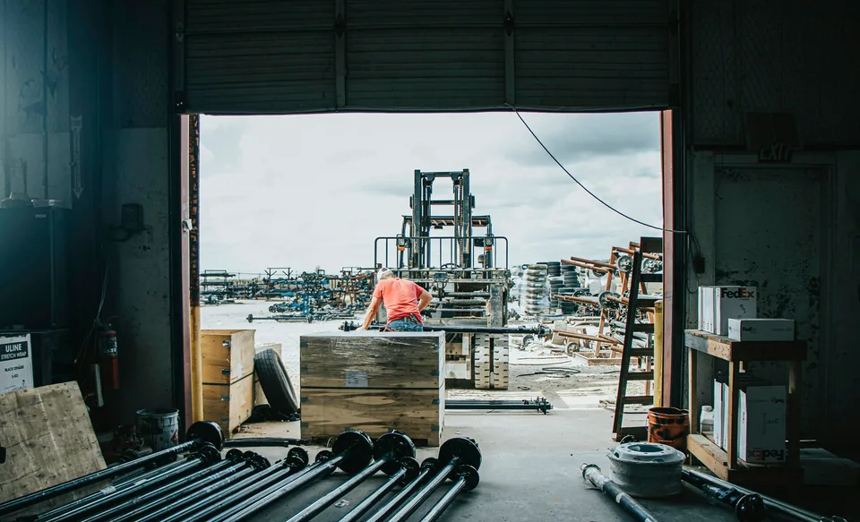 Looking out through a trailer shop's open bay door to the yard, where a worker bends over a crate by a forklift; trailer axles lie on the shop floor inside.
